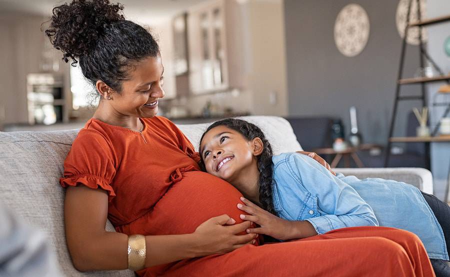 A pregnant woman in an orange dress sitting on the couch with her daughter and smiling.
